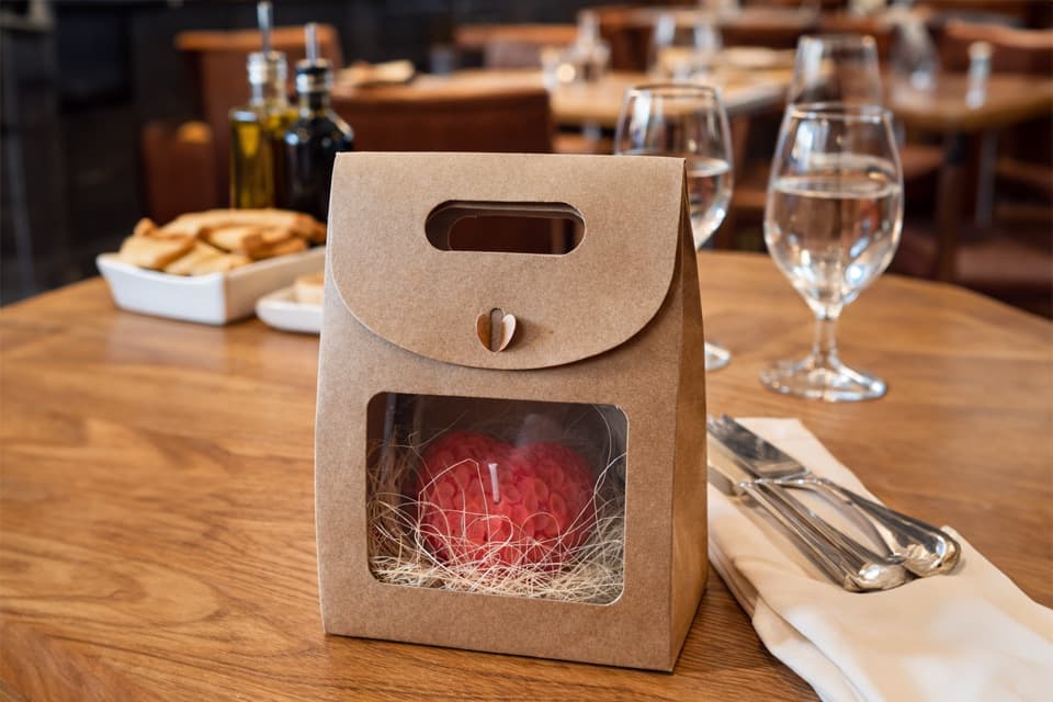 Bougie moulée rouge présentée dans un coffret cadeau en kraft avec fenêtre transparente, posée sur une table de restaurant élégamment dressée