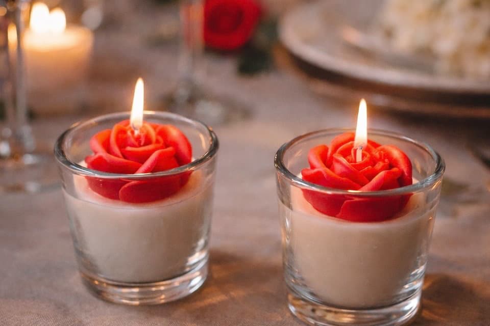 Deux bougies artisanales en verre, avec une fleur rouge en cire au centre et la flamme allumée, dans une ambiance douce et chaleureuse